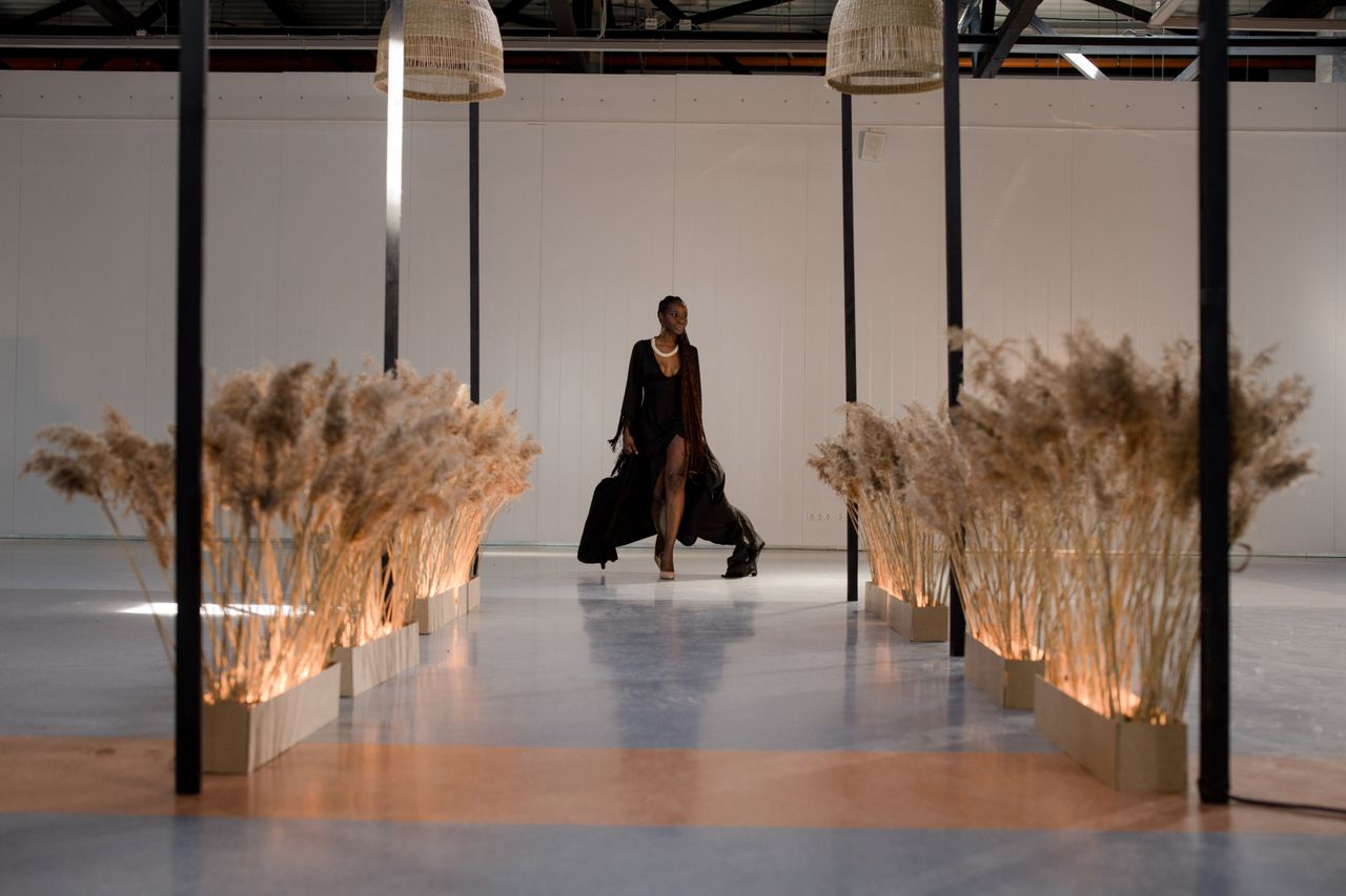 Graceful model in a black dress walking on a runway indoors, surrounded by minimalist decor.
